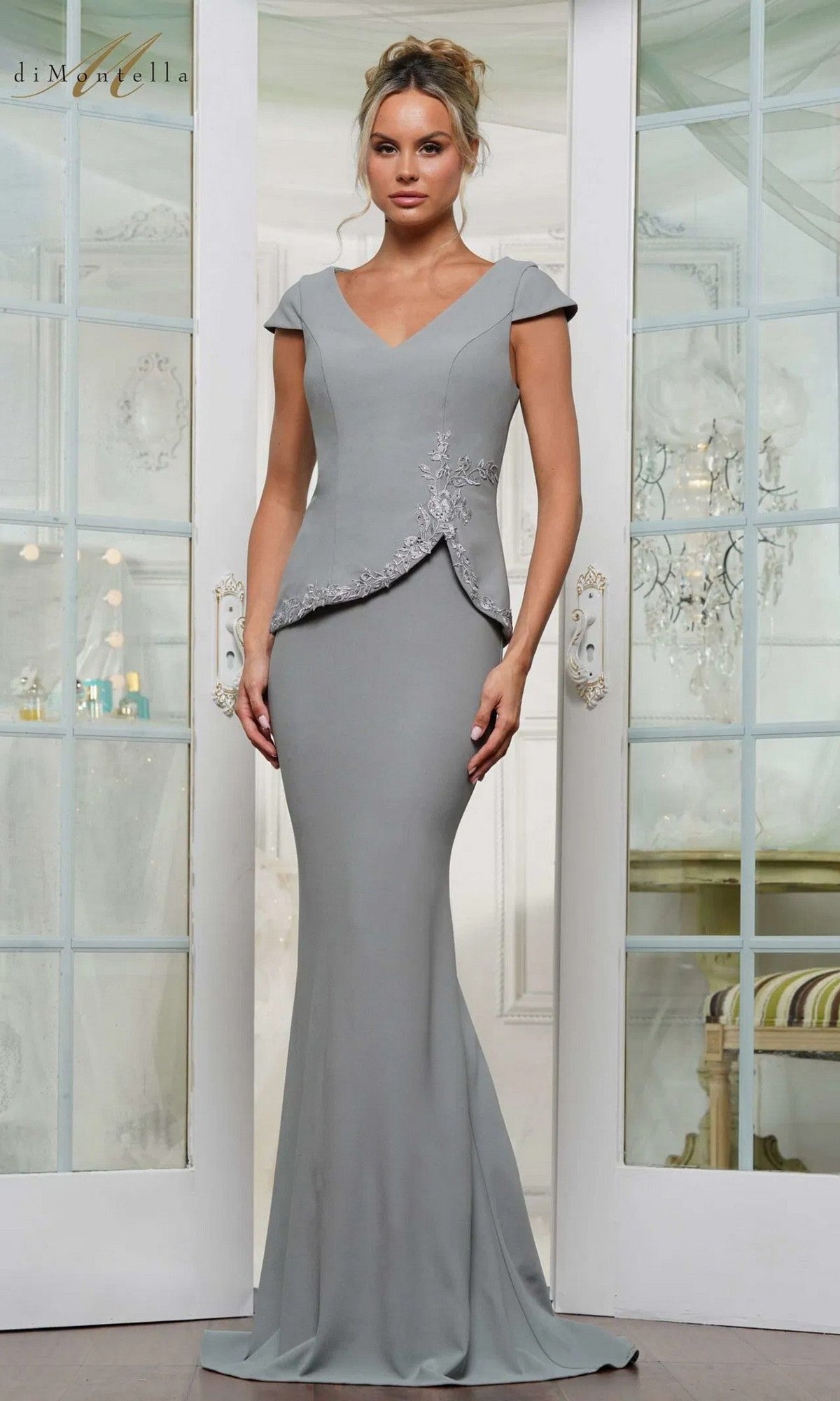 A woman stands indoors wearing the Long Formal Dress: Rina Di Montella RD3239 in Seaglass, a fitted stretch crepe gown with short sleeves, embroidered waist detail, and a graceful mermaid skirt.